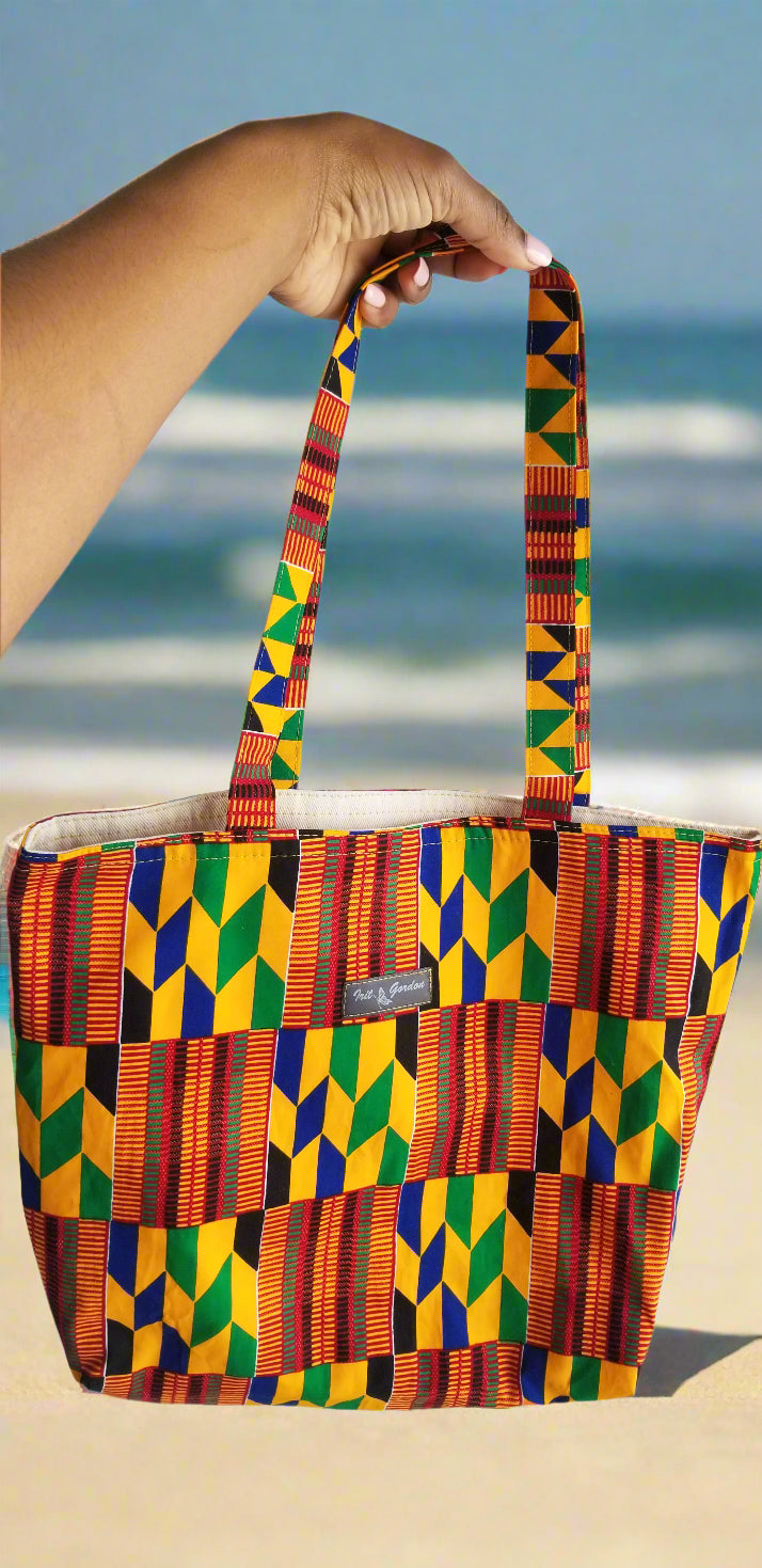 Vibrant Kente print tote bag with geometric patterns by Scope and Flow, styled against a blurred beach background.
